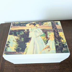 May include: A rectangular white box featuring a printed reproduction of an impressionist painting on its lid. The artwork depicts a person in a light blue dress and hat standing near a child in a blue dress beside a dark tree trunk.