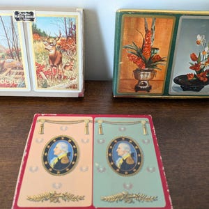 May include: Three rectangular playing card boxes. One box features a deer and a pheasant with the text PINOCHLE. Another box displays two floral arrangements in vases. The third box shows two oval portraits of George Washington in uniform.