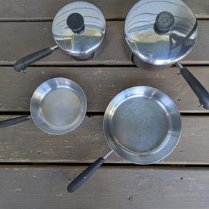 May include: A set of four stainless steel cookware pieces, including two pots with lids and two frying pans. The pots have black handles and lids with black knobs. The pans also have black handles. The cookware is arranged on a wooden surface.