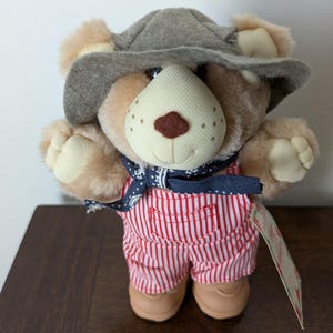 May include: A plush teddy bear wearing a gray hat, a blue bandana, and red and white striped overalls. The bear has a beige snout and paws, and is wearing tan shoes. A tag is attached to the overalls.