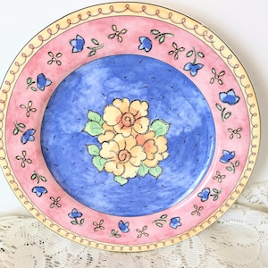 May include: A round ceramic plate featuring a central cluster of yellow flowers with green leaves on a blue background. The rim is pink with a repeating pattern of small blue flowers and green sprigs, finished with a thin yellow edge and black line.