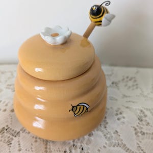 May include: A decorative honey pot with a lid, painted in a light yellow color. The lid is topped with a white flower. A small bee is attached to the lid. Another bee is painted on the side of the pot. The pot is sitting on a white lace surface.