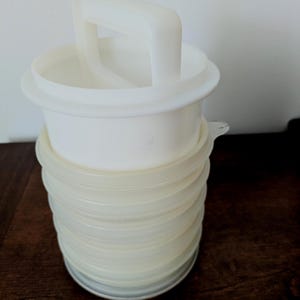 May include: A white plastic kitchen tool featuring a cylindrical, collapsible accordion-style base and a top section with a vertical handle. The structure consists of multiple stacked rings that allow for height adjustment, finished in a smooth, opaque white.