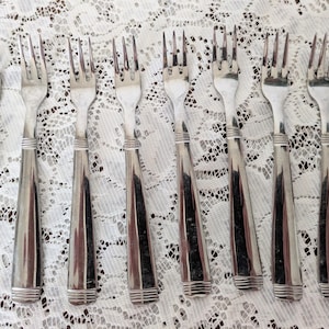May include: A collection of eight silver forks with a classic design. Each fork features a three-tine head and a long, tapered handle with a decorative band near the top and bottom. The forks are arranged on a white lace tablecloth.