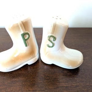 May include: A pair of ceramic salt and pepper shakers shaped like cowboy boots. One boot has a green "P" and the other has a green "S". The boots are off-white with tan accents and small holes at the top.