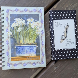 May include: Two notebooks on a wooden surface. One is a spiral-bound notebook with a floral design featuring white flowers in a blue pot. The other is a small, black notebook with white polka dots and the text "Chelsea" and "The less the shoe the more intriguing the View."