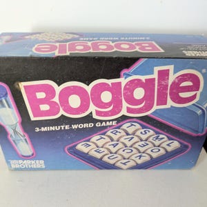 May include: A vintage Boggle word game box. The box features the word "Boggle" in large pink letters, with a blue and black background. The box also includes a 3-minute timer and a tray of lettered dice.