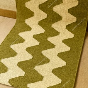 May include: A green and cream-colored rug with a zig-zag pattern. The rug is made of a soft material and has a textured surface. The zig-zag design is a repeating pattern of cream-colored chevrons against a green background.