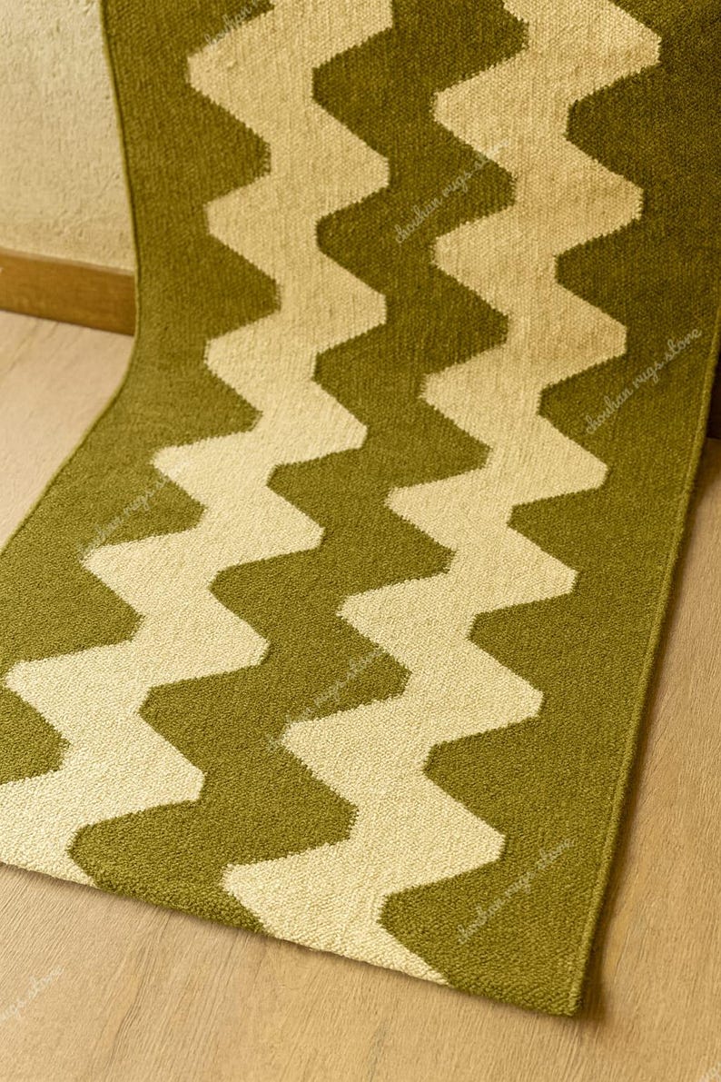 May include: A green and cream-colored rug with a zig-zag pattern. The rug is made of a soft material and has a textured surface. The zig-zag design is a repeating pattern of cream-colored chevrons against a green background.