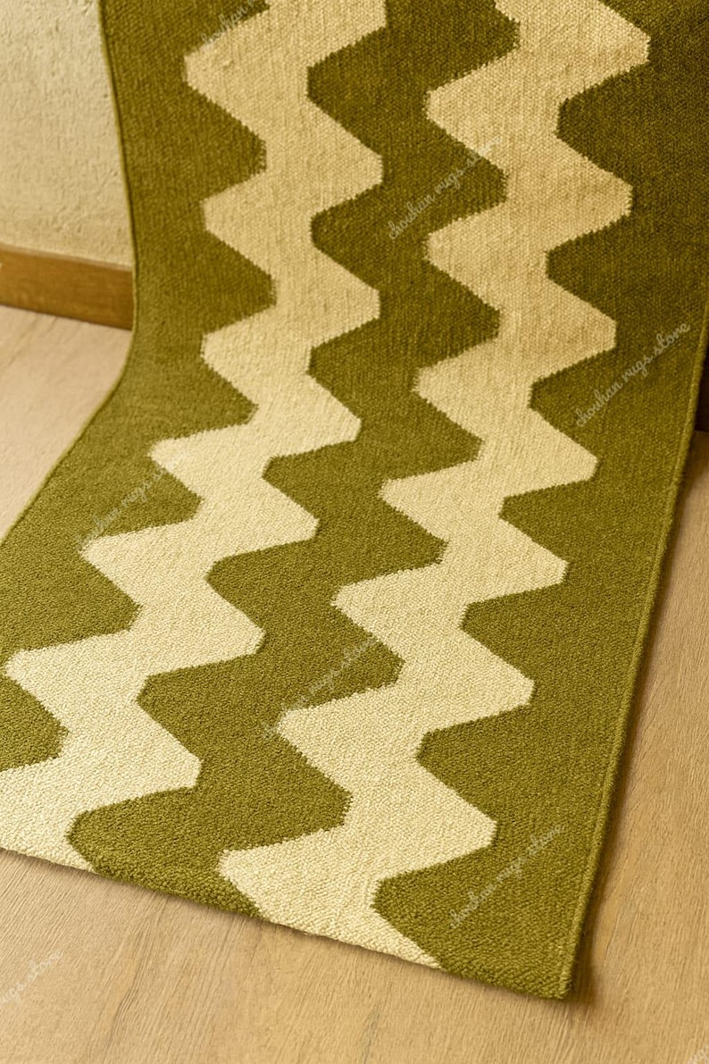 May include: A green and cream-colored rug with a zig-zag pattern. The rug is made of a soft material and has a textured surface. The zig-zag design is a repeating pattern that runs the length of the rug.