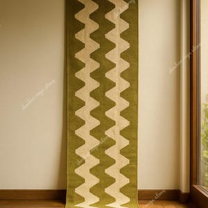 May include: A long, olive green and beige zig-zag patterned rug hanging on a wall. The rug is made of a woven material and is displayed vertically. The rug is approximately 8 feet long.