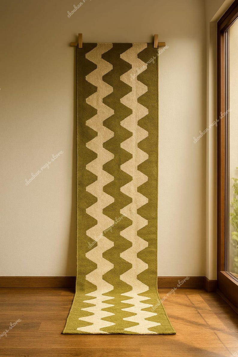 May include: A long, olive green and beige zig-zag patterned rug hanging on a wall. The rug is made of a woven material and is displayed vertically. The rug is approximately 8 feet long.