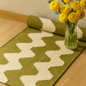 May include: A green and cream-colored zigzag patterned rug is partially rolled up, with a matching rug laid out. A vase of yellow flowers sits nearby. The rug is made of a soft, textured material.