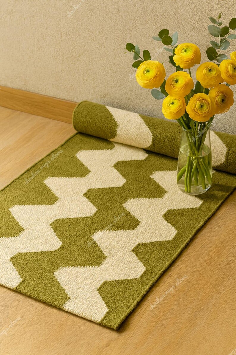 May include: A green and cream-colored zigzag patterned rug is partially rolled up, with a matching rug laid out. A vase of yellow flowers sits nearby. The rug is made of a soft, textured material.