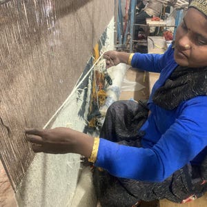 May include: A person weaving a rug on a large loom. The rug is a mix of brown and white threads. The person is wearing a blue shirt and a black scarf. Gold bracelets adorn their wrists.