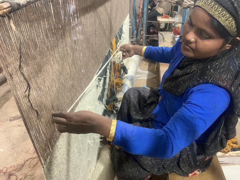 May include: A person weaving a rug on a large loom. The rug is a mix of brown and white threads. The person is wearing a blue shirt and a black scarf. Gold bracelets adorn their wrists.