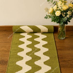 May include: A green and white zigzag patterned runner rug is partially rolled up on a wooden floor. A vase of yellow roses sits to the right. The rug's design is a repeating series of white zigzags on a green background.