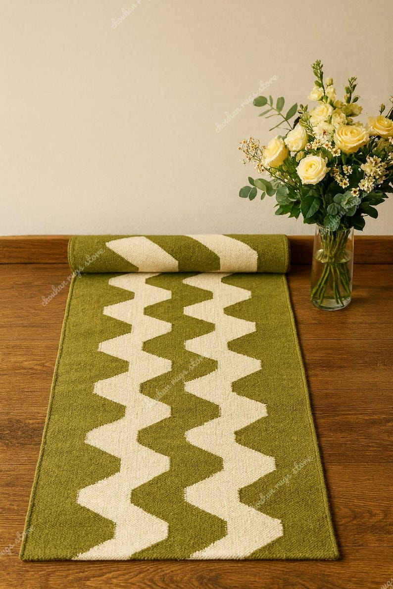 May include: A green and white zigzag patterned runner rug is partially rolled up on a wooden floor. A vase of yellow roses sits to the right. The rug's design is a repeating series of white zigzags on a green background.
