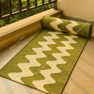 May include: A green and cream-colored rug with a zigzag pattern. The rug is rectangular and partially rolled up, with the rolled portion resting on the floor. The rug is made of a soft material and is suitable for indoor or outdoor use.