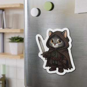 Pode incluir: Um íman de desenho animado com um gato cinzento com uma capa com capuz castanha e uma espada prateada. O gato tem olhos amarelos e está vestido como um guerreiro medieval.