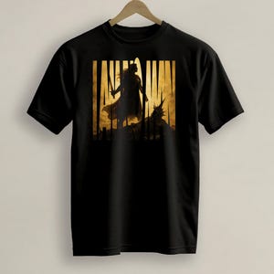 Pode incluir: T-shirt preta com um design de silhueta dourada e preta de uma figura com uma espada, em pé em frente a uma criatura. O fundo tem linhas verticais. A t-shirt está pendurada num cabide de madeira.