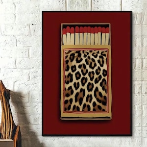 May include: A framed artwork depicting a matchbox. The matchbox design features a leopard print pattern and red-tipped matches. The artwork is set against a deep red background and framed in black.