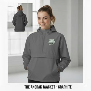 May include: Graphite anorak jacket with a hood and a front pocket. The jacket has a half-zip closure and the text "YOUR DESIGN HERE" on the chest. The jacket is shown on a person. The text "THE ANORAK JAACKET - GRAPHITE" is at the bottom.