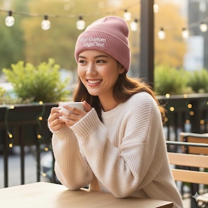 May include: A light pink beanie with the text "Your Custom Logo/Text" in white. The beanie is worn by a person wearing a cream-colored sweater. The person is holding a white mug and smiling.