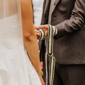 May include: A couple exchanging vows during a wedding ceremony. The groom is wearing a dark suit and holding a white, green and black braided cord. The bride is wearing a white dress and veil.