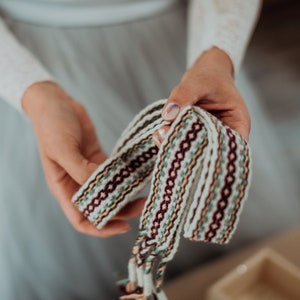 May include: A woven belt with a repeating pattern of burgundy, teal, and gold on a cream background. The belt is held up in front of a light blue background.