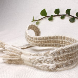 May include: A handwoven belt in natural tones of beige and light brown. The belt features a textured weave with a braided section and fringed ends. A sprig of green leaves is in the background.