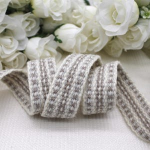 May include: A patterned ribbon in shades of grey and brown, curled on a white surface. White roses are in the background. The ribbon's design features a repeating pattern, suitable for crafts or decorative purposes.