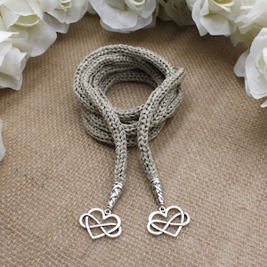 May include: A beige knitted cord with two infinity heart charms. The cord is coiled and the charms are hanging down.