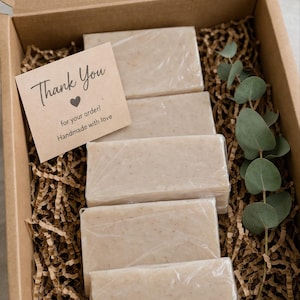Può includere: Una scatola di cartone piena di cinque saponette avvolte, adagiate su carta marrone. Nella scatola sono presenti anche un piccolo biglietto con la scritta "Thank You" e un rametto di eucalipto. Le saponette sono beige chiaro.