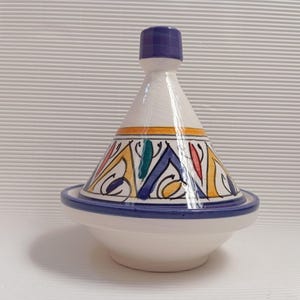 Pode incluir: Uma panela de tagine de cerâmica branca com tampa cónica e base redonda. A tampa é decorada com padrões geométricos coloridos e um botão azul. A base e a borda são azuis, e a panela é projetada para cozinhar e servir.