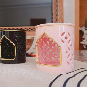 May include: Two cylindrical ceramic oil burners, one in glossy black and one in soft pastel pink. Each burner features a gold-rimmed arched opening and intricate laser-cut geometric patterns on the sides, designed for holding a tea light candle.