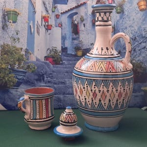 Peut inclure: Un ensemble de pichet, tasse et couvercle en céramique. Le pichet est orné de motifs géométriques bleus, rouges, jaunes et blancs. La tasse présente des rayures verticales, et le couvercle est arrondi. L'ensemble est posé sur une surface verte.