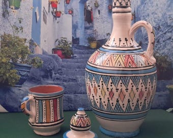 May include: A ceramic pitcher, mug, and lid set. The pitcher has a handle and is decorated with geometric patterns in blue, red, yellow, and white. The mug has vertical stripes, and the lid has a rounded top. The set is on a green surface.