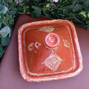 Peut inclure: Un couvercle de tajine carré, de couleur terre cuite, avec un motif décoratif et orné en or. Le couvercle comporte un bouton central et est entouré d'une bordure détaillée. Le couvercle de tajine est un ustensile de cuisine traditionnel.