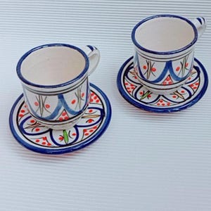 Peut inclure: Deux tasses à expresso et soucoupes en céramique assorties. Les tasses et les soucoupes sont blanches avec des motifs décoratifs bleus, rouges et verts. Les tasses ont de petites anses et sont posées sur les soucoupes.
