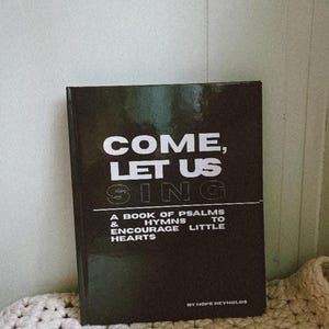 May include: A black book titled "COME, LET US SING" with white lettering. The book is a collection of psalms and hymns to encourage little hearts. The book is resting on a cream-colored crocheted blanket.