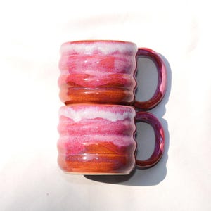 May include: Two stacked ceramic mugs featuring a ribbed, wavy texture. The glaze transitions from a deep reddish-brown at the base to a vibrant magenta in the middle, topped with a creamy white drip effect along the rim and a curved handle.