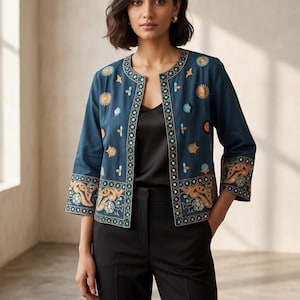 Op de afbeelding: Een teal jasje met ingewikkelde borduursels in oranje, geel en blauw. Het jasje heeft een ronde halslijn en driekwart mouwen. Het model draagt een zwarte top en een zwarte broek. Het ontwerp van het jasje heeft bloemen en geometrische patronen.