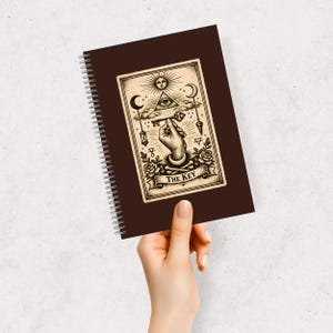 May include: A brown spiral-bound notebook with a tarot card illustration. The card shows a hand holding a key, with celestial symbols. The text "THE KEY" is on a banner. The notebook is held in a hand.