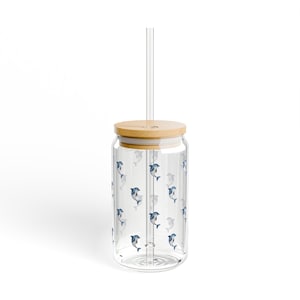 May include: A clear glass tumbler with a bamboo lid and a clear straw. The tumbler is adorned with a repeating pattern of blue and gray dolphins. The tumbler is designed for cold beverages.