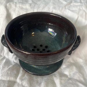 May include: A handcrafted ceramic colander with a dark, glossy glaze. The rim is a deep brown, fading into a teal-green interior. It has small drainage holes and two curved handles. A functional kitchen item.