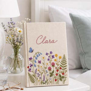 Op de afbeelding: Een crèmekleurig, met stof bekleed boek met de naam Clara geborduurd in roze schrijfletters. De kaft is versierd met kleurrijke geborduurde wilde bloemen, groene varens, kleine rode aardbeien en een enkele blauwe vlinder.