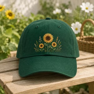 Könnte beinhalten: Eine dunkelwaldgrüne Baseballkappe mit einer Stickerei von drei gelben Sonnenblumen mit braunen Mitten, umgeben von grünen Stielen, Blättern und dünnen Weizenhalmen, die mittig auf der Vorderseite der Kappe platziert ist.