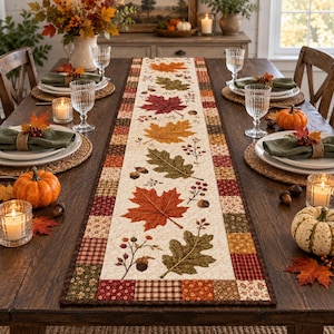 Könnte beinhalten: Ein rechteckiger, gesteppter Tischläufer mit einem cremefarbenen Mittelteil, der mit einem wiederkehrenden Muster aus Herbstblättern, Eicheln und Beeren verziert ist, umrandet von einem Patchwork-Design aus kleinen Quadraten in Braun, Orange und Grün.