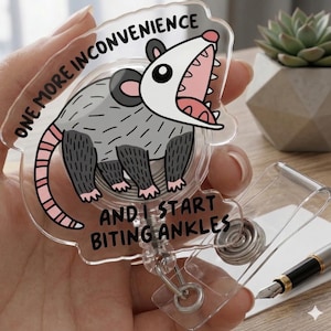 May include: A clear acrylic badge reel featuring a cartoon opossum with an open mouth. The opossum is gray with black and pink accents. Text on the badge reel reads "ONE MORE INCONVENIENCE AND I START BITING ANKLES."